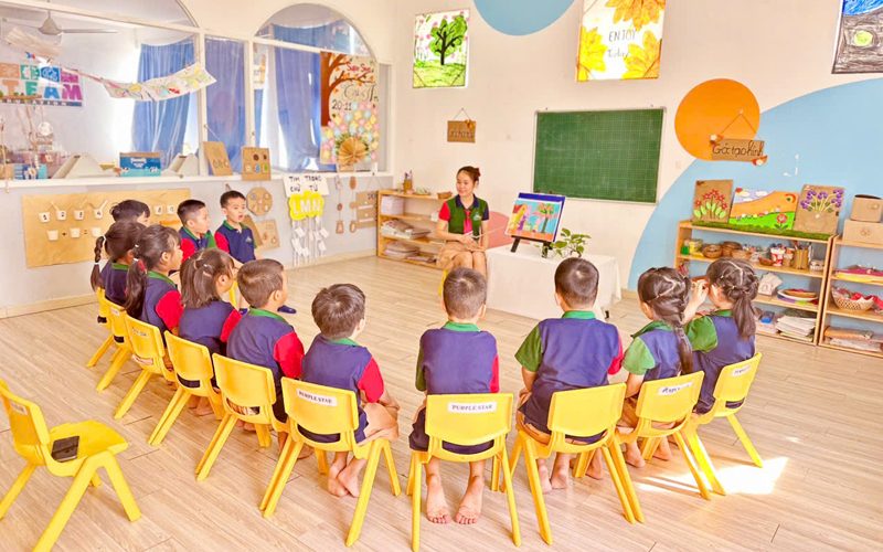 Cả lớp ngồi ghế vàng thành vòng tròn nghe cô kể chuyện trong phòng học sáng màu