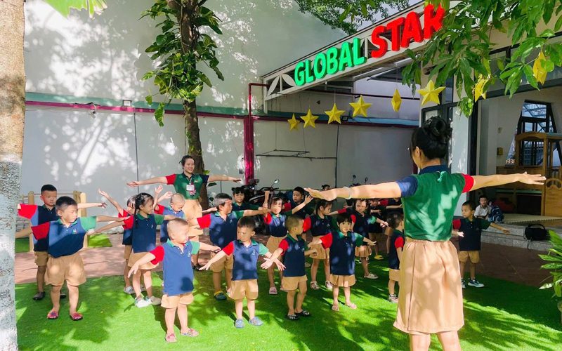 Cô và các bé dang tay tập thể dục trên sân cỏ trước cổng trường
